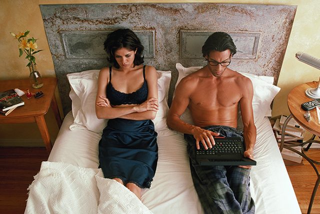 Couple in bed, man using laptop, woman with displeased expression